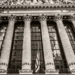 Front facade of the New York Stock Exchange with tall Corinthian columns and American flags on either side.