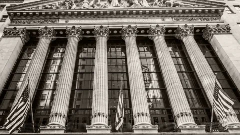 Front facade of the New York Stock Exchange with tall Corinthian columns and American flags on either side.