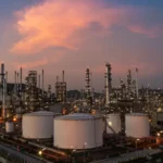 Industrial refinery complex at dusk with tall distillation columns, piping, and large white storage tanks, hills in the background.
