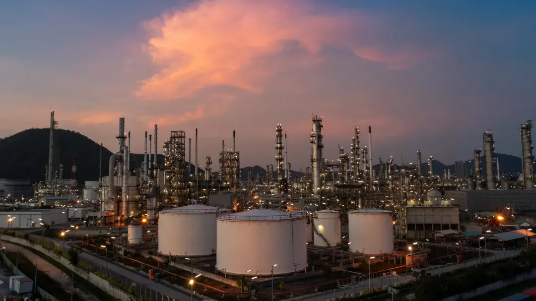 Industrial refinery complex at dusk with tall distillation columns, piping, and large white storage tanks, hills in the background.