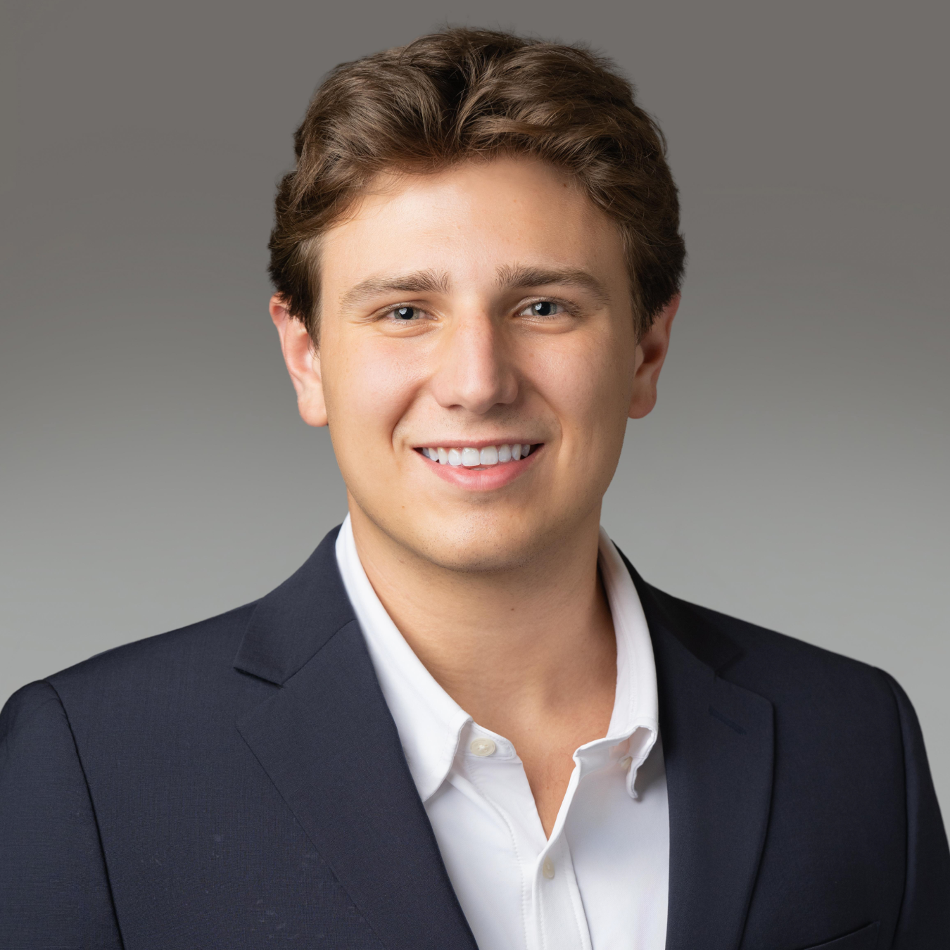 Professional headshot of a smiling young man in a navy blazer and white shirt, against a gray gradient studio backdrop.