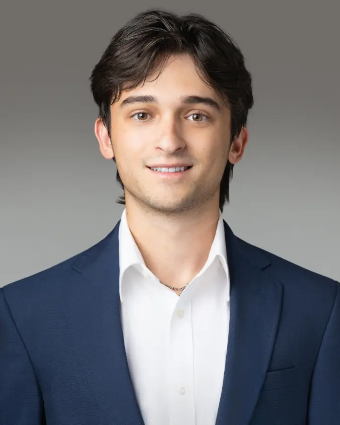 Young man in a navy blazer and white shirt smiling at the camera against a gray background.