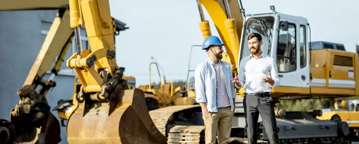 Two professionals stand and talk beside a yellow excavator on a construction site.