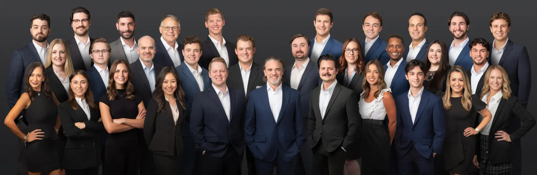 Group of 25 professionals in business attire posing together for a team photo against a dark backdrop, conveying a corporate team or company leadership image.