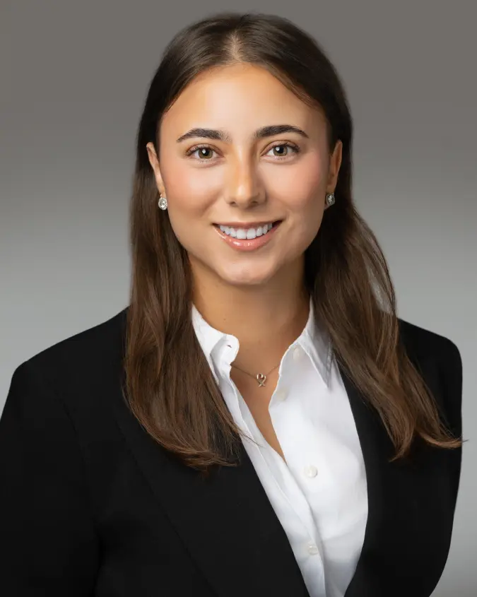 Smiling professional headshot of a woman with long brown hair, wearing a black blazer and white blouse against a gray gradient background.